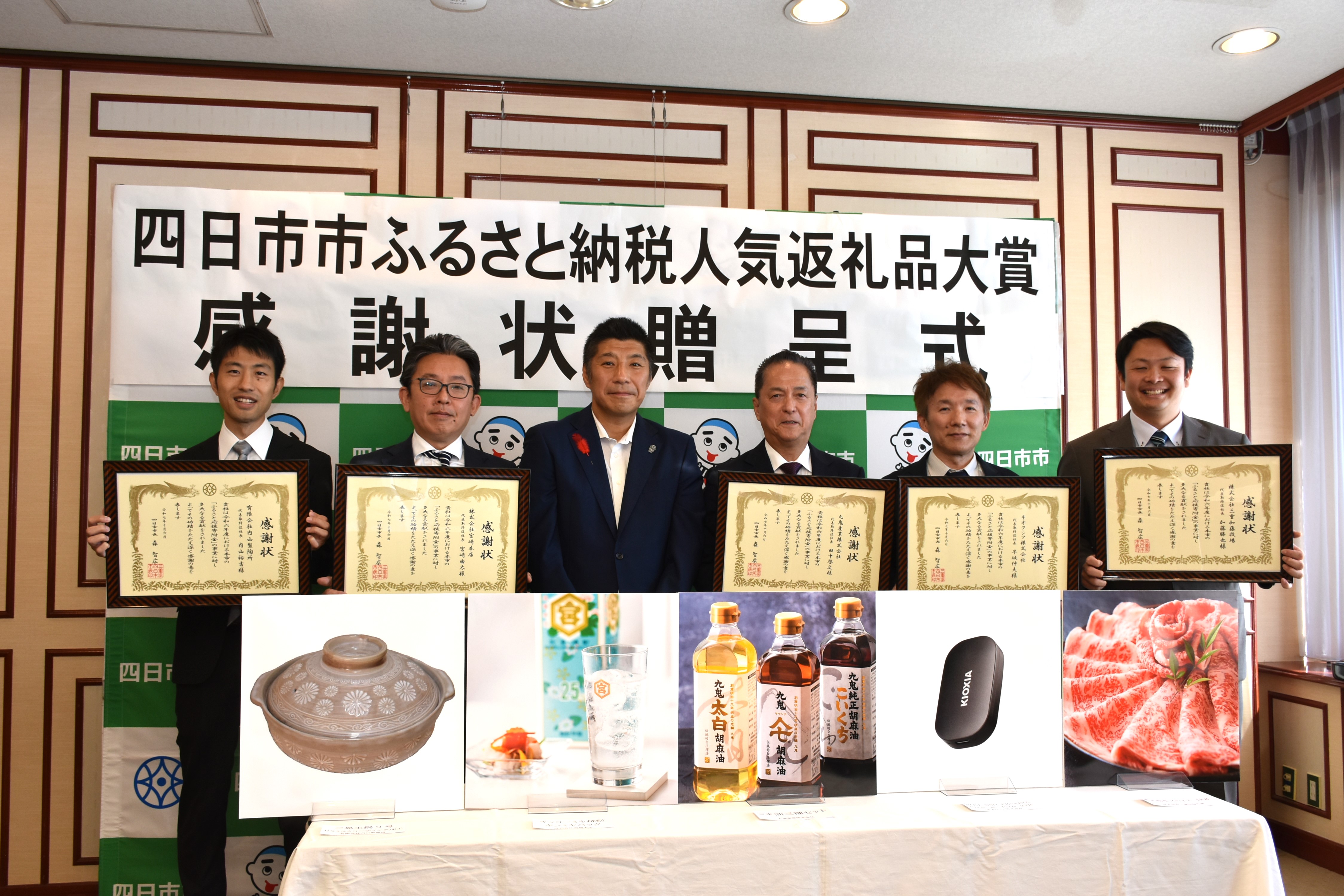 感謝状を持つ受贈者と市長、前には返礼品のパネルが並ぶ